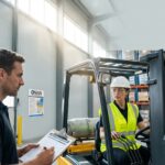 Warehouse forklift operator training with trainer holding a clipboard and a visible OSHA compliance checklist on the wall